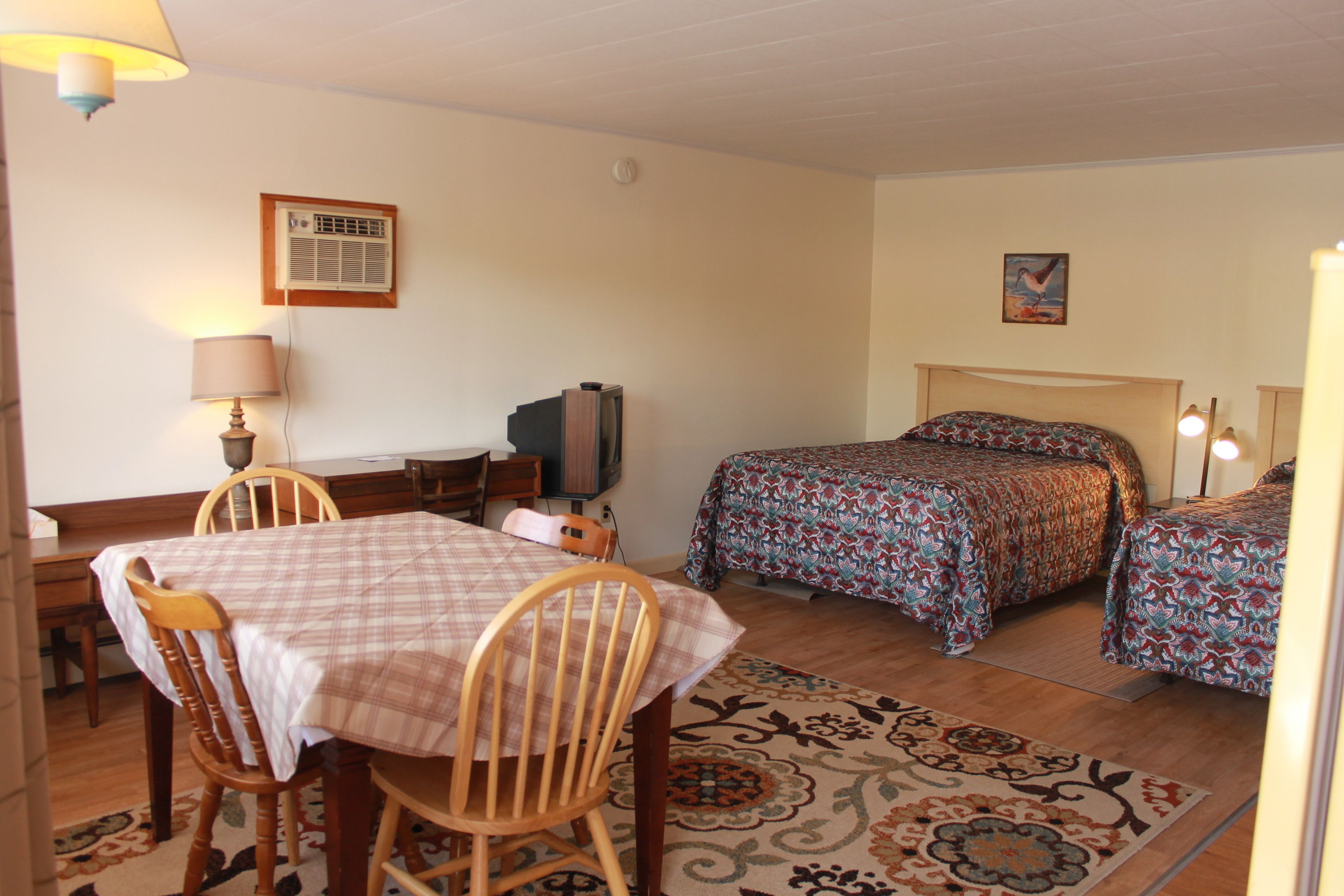 A room with a dining table, two beds with patterned bedding, a television, and a wall-mounted air conditioner.