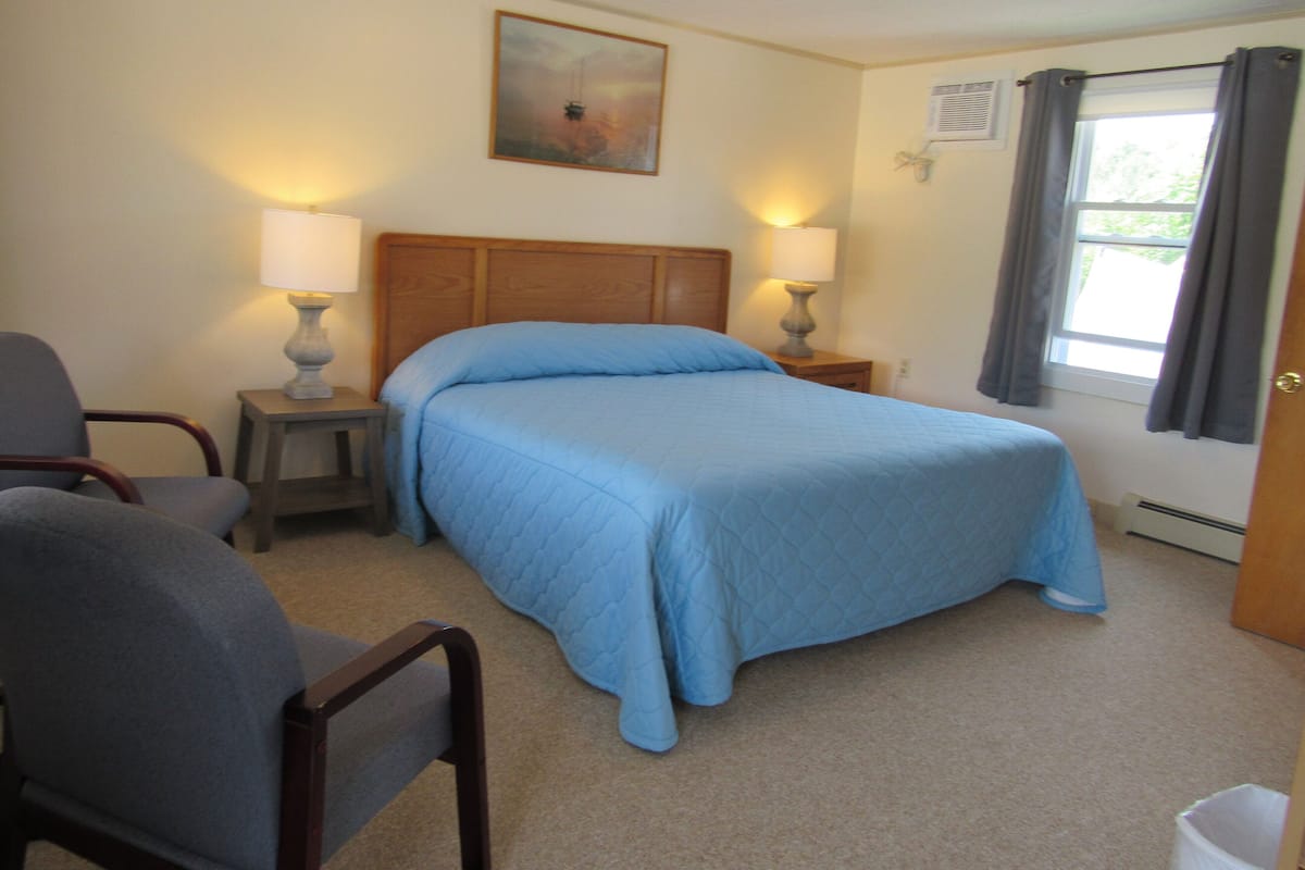 A bedroom with a bed, two bedside lamps, a chair, a painting on the wall, and a window with curtains.
