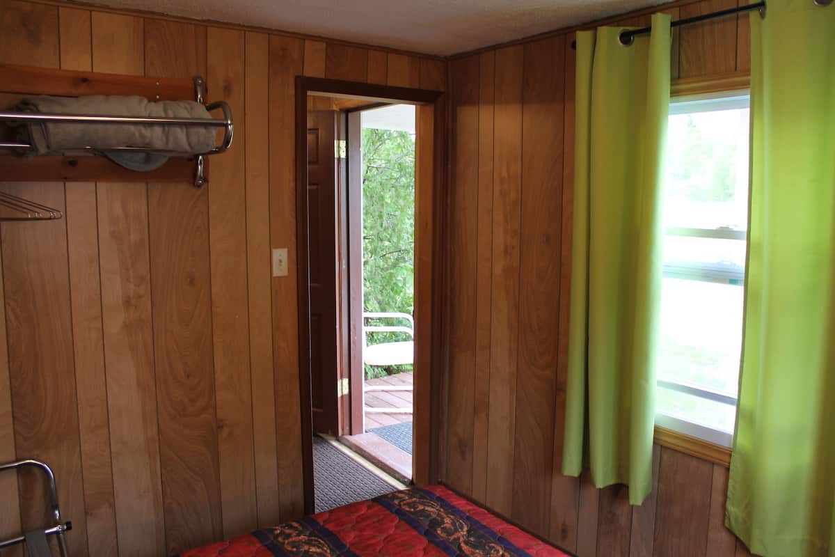 A room with wooden paneling, a bed with a patterned bedspread, a metal rack, and a window with green curtains.