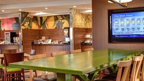A hotel dining area with a large green table and wooden chairs, a buffet counter, and a digital screen displaying weather information.