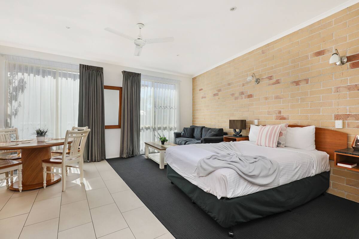 A modern hotel room with a large bed, a dining table with chairs, a sofa, and a brick accent wall.