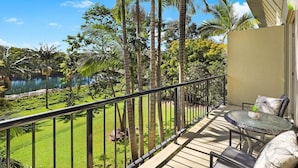 A balcony with a glass table and chairs, overlooking a lush garden and a body of water.