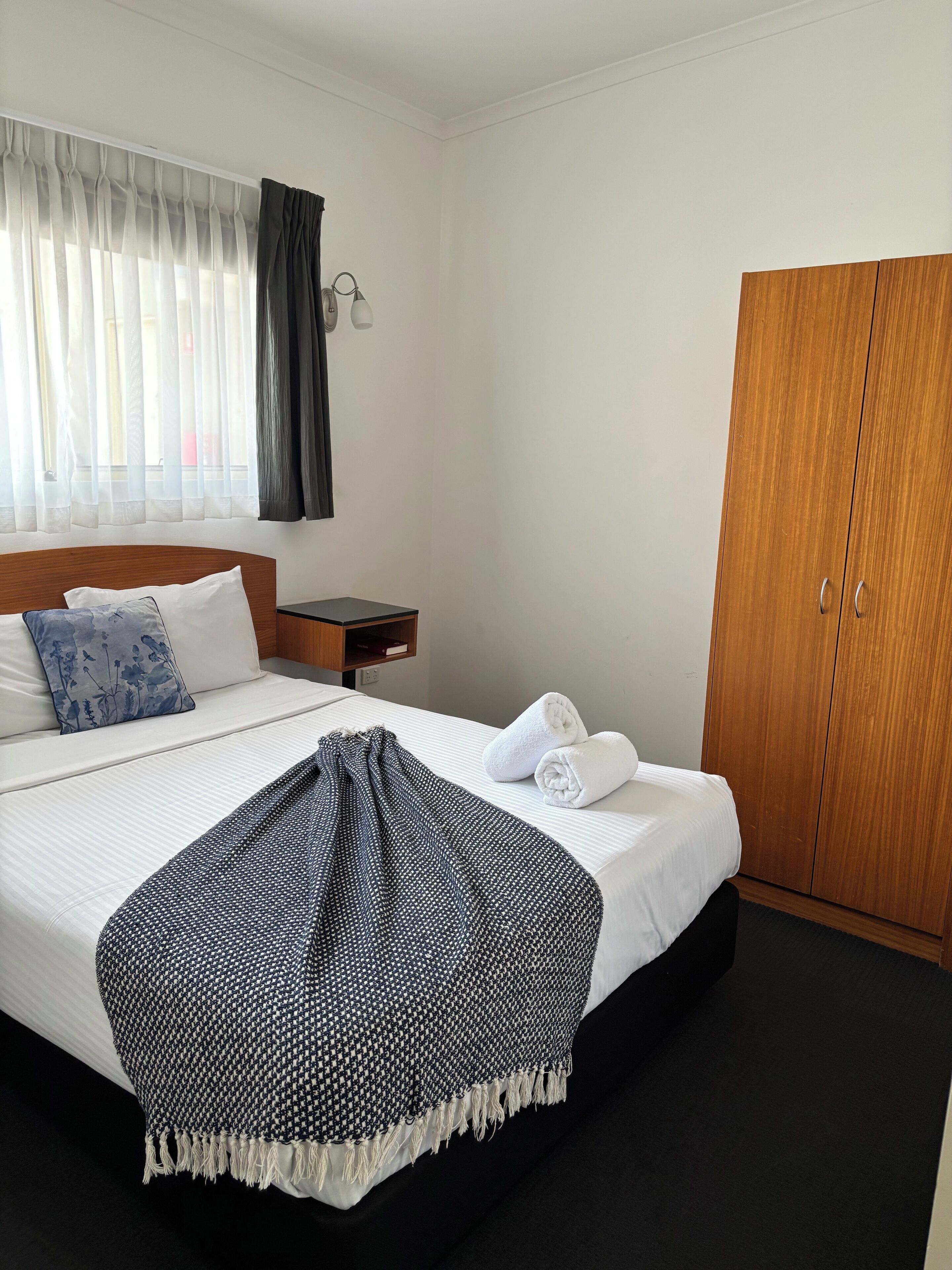 A hotel room with a bed, a wooden wardrobe, a nightstand, and a window with curtains.