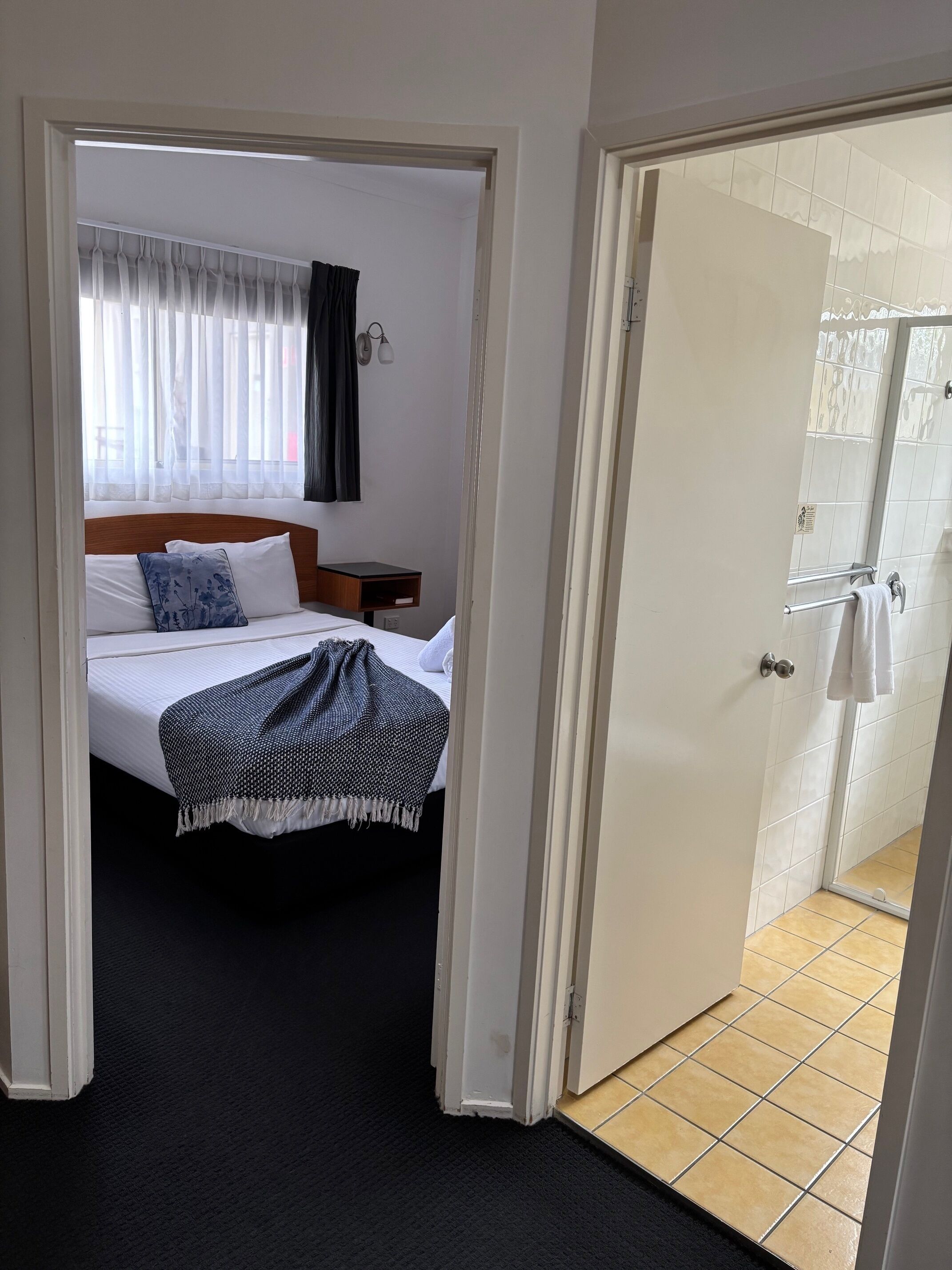 A hotel room with a bed, a nightstand, a window with curtains, a bathroom with a shower, and a tiled floor.