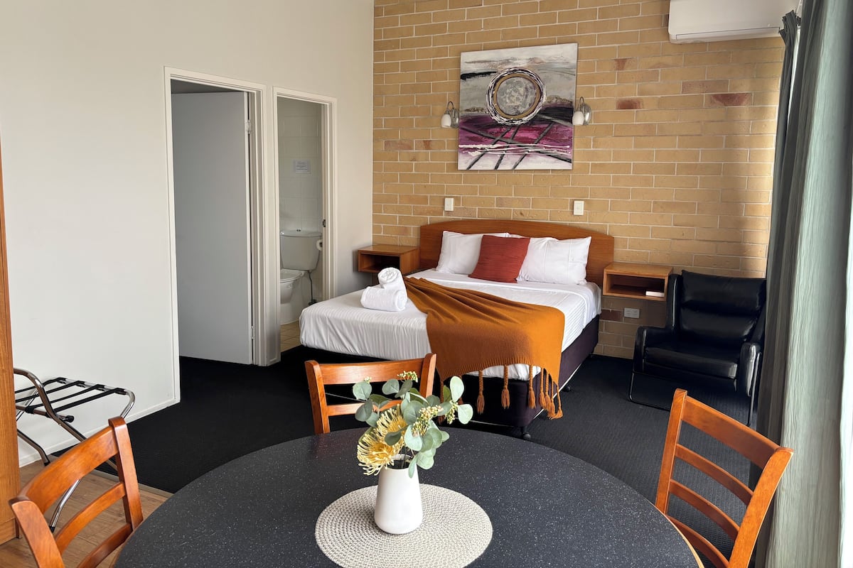 A hotel room with a bed, a bedside table, a chair, a dining table with chairs, a vase with flowers, and a painting on the wall.