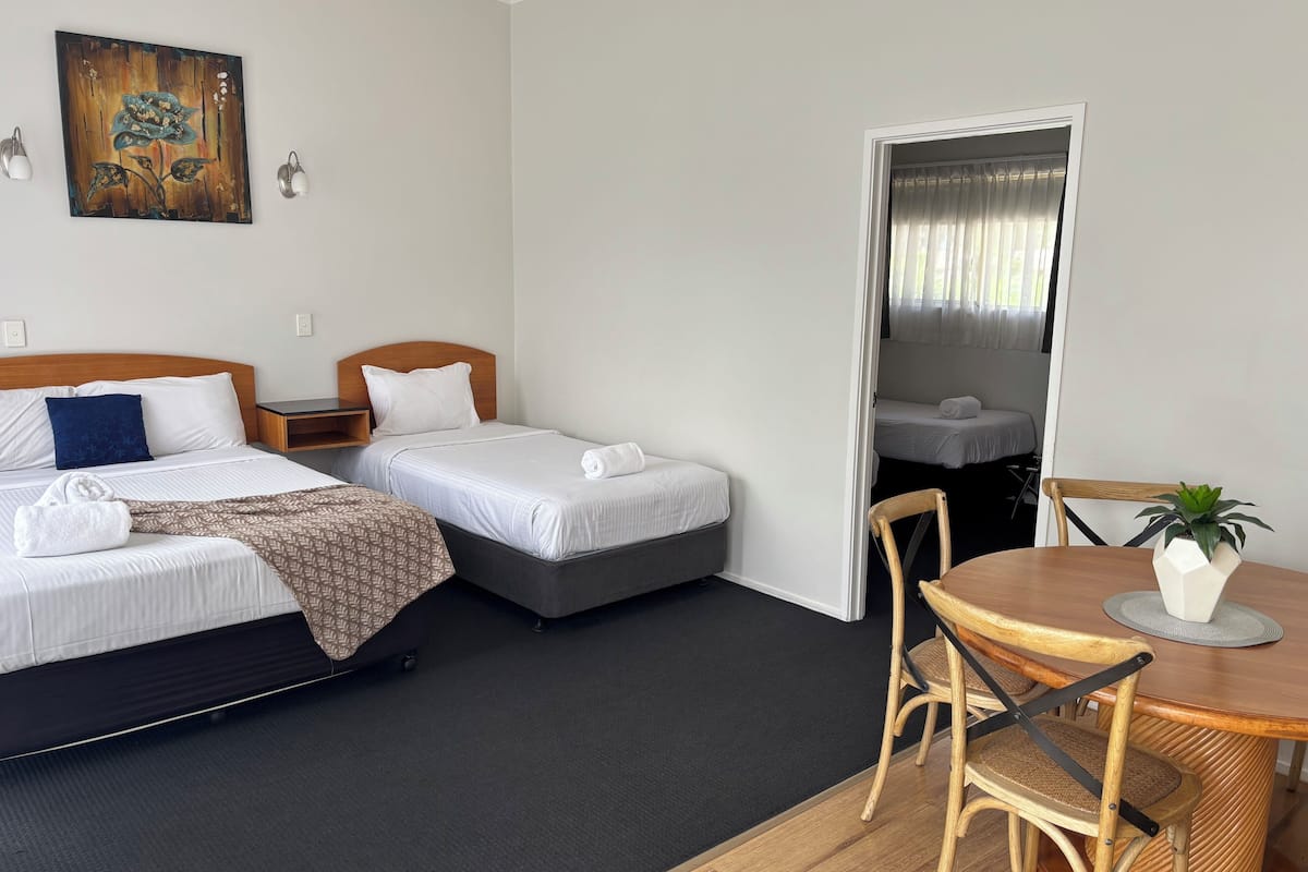 A hotel room with two beds, a wooden headboard, a small table with a chair, and a potted plant.