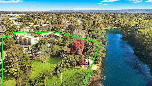 An aerial view of a residential area with houses, a river, and a green space with palm trees.