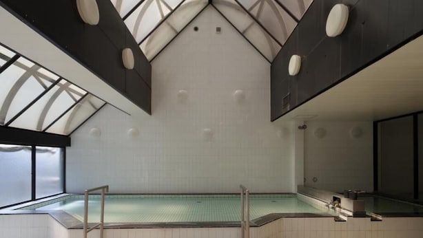 A large indoor swimming pool with a high ceiling and skylights, surrounded by white tiled walls and a staircase leading to the pool area.