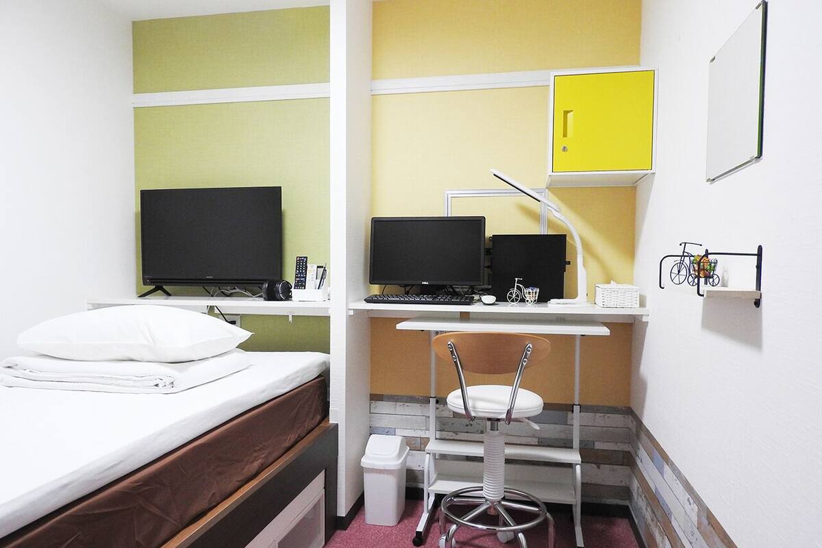 A compact hotel room with a bed, two desks with computers, a chair, and a small trash bin.