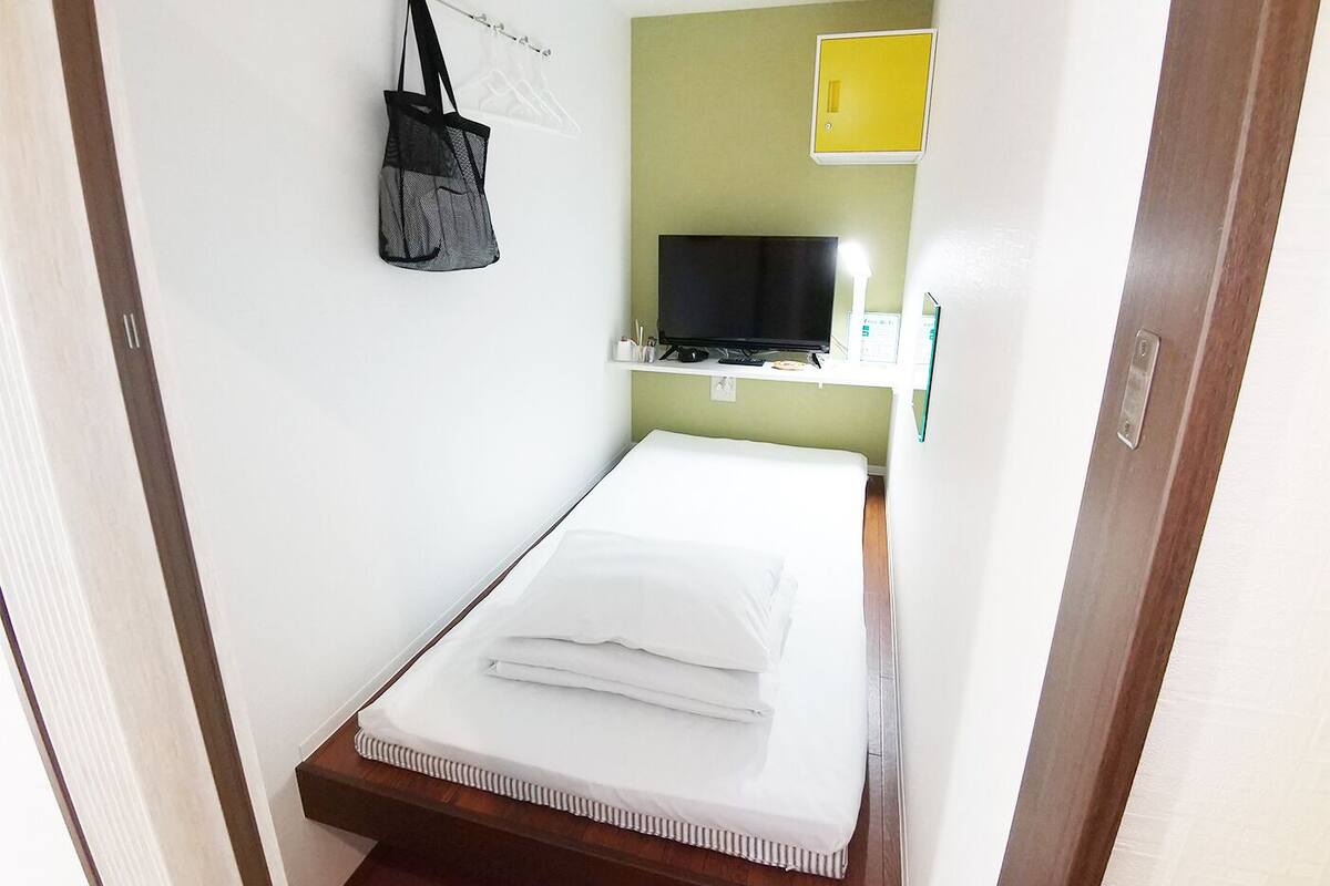 A small, neatly made bed with white linens in a compact room with a green wall and a yellow light fixture.