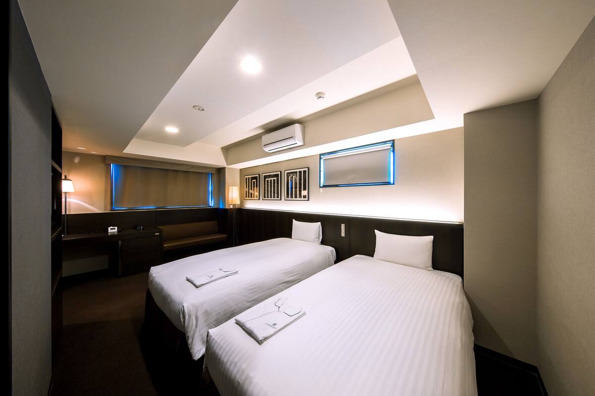 Une chambre d’hôtel avec deux lits simples, un bureau, une chaise et une grande fenêtre.