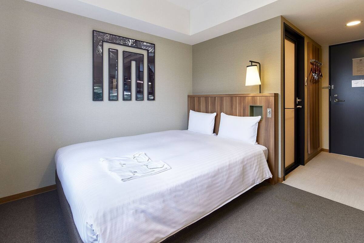 Une chambre d’hôtel comprenant un lit, une table de chevet, une lampe fixée au mur et une porte avec un porte-manteau.