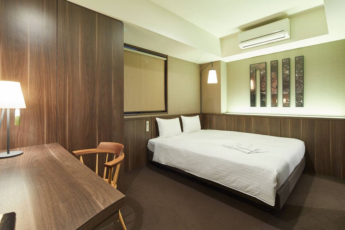 Une chambre d’hôtel avec un bureau en bois, un lit, une chaise, un climatiseur fixé au mur et une fenêtre.