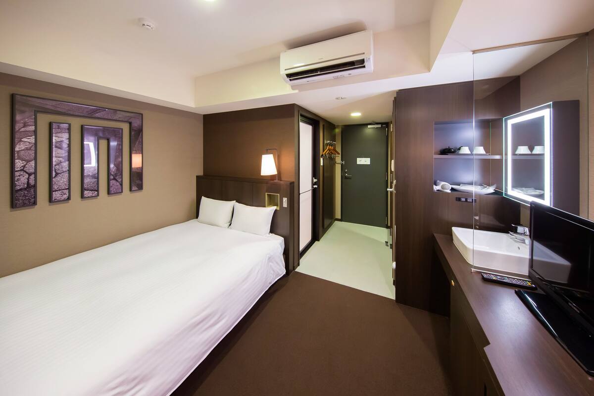 Une chambre d’hôtel comprenant un lit, une table de chevet avec une lampe, un miroir, une télévision et une salle de bain avec un lavabo et un miroir.