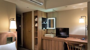 A hotel room with a bed, a desk with a TV, a chair, a mirror, and a bathroom with a sink and towel storage.