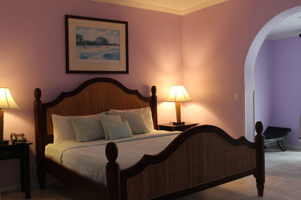 A neatly made bed with a headboard, two bedside lamps, a framed picture on the wall, and a bench in the adjacent room.