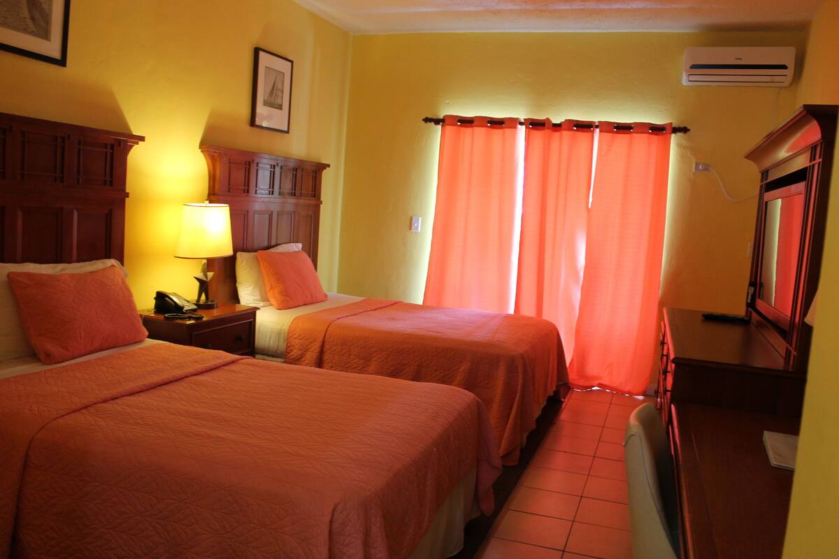 A hotel room with two beds, a nightstand with a lamp, a dresser, and an air conditioning unit.