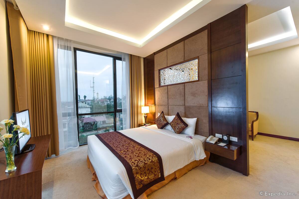 A hotel room with a large bed, a desk with a lamp, a phone, and a view of the city through a large window.
