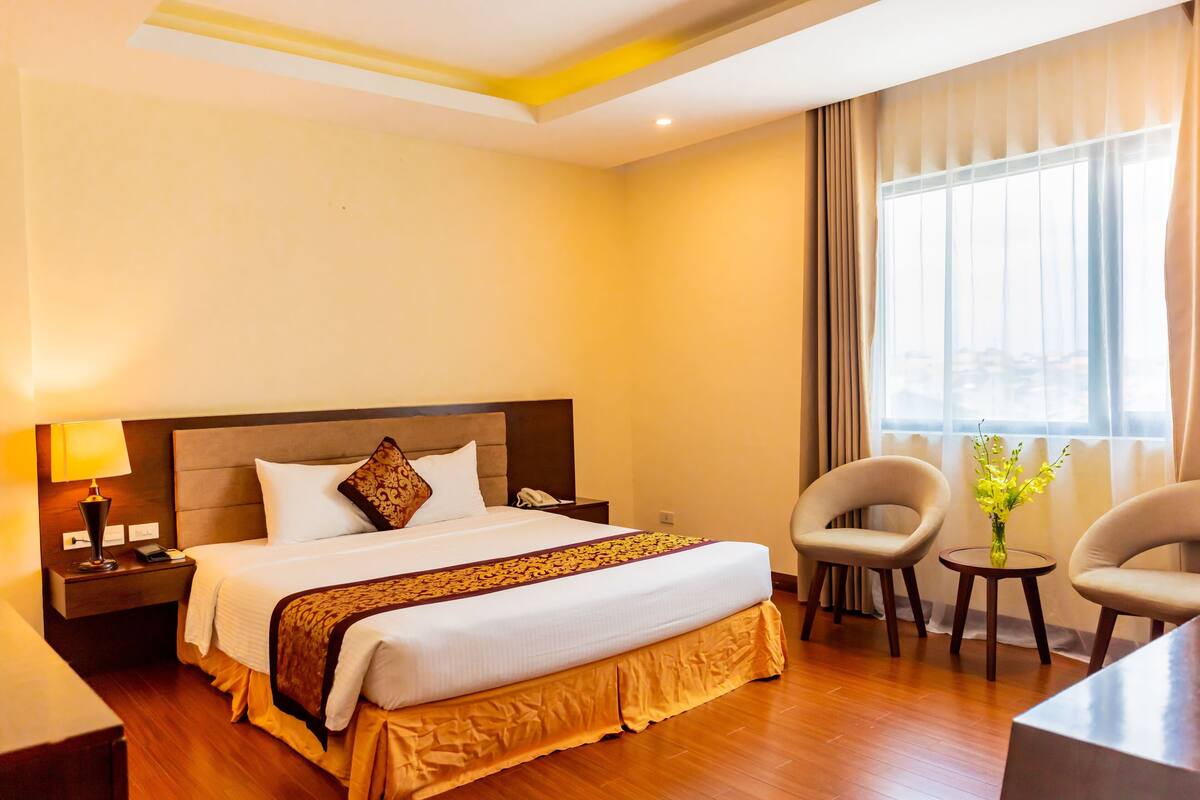 A hotel room with a large bed, bedside tables, a lamp, two chairs, a small table with a vase, and a window with curtains.