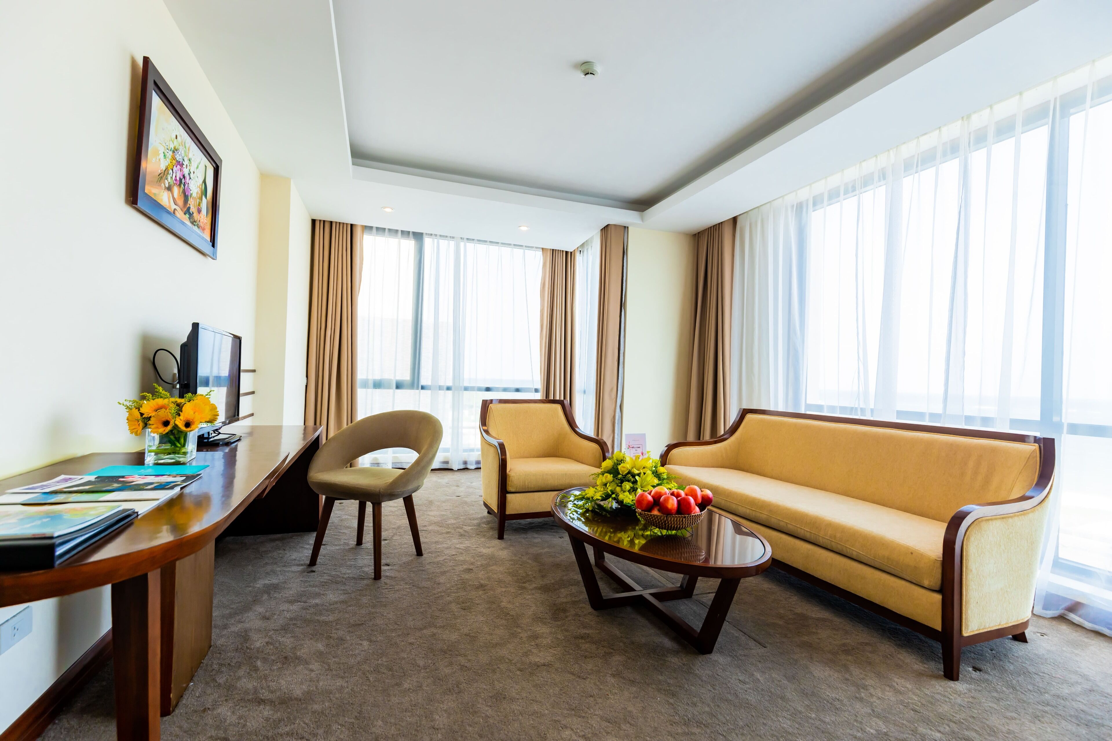 Habitación de hotel con sofá, sillón, mesa de centro y escritorio con computadora y un florero.