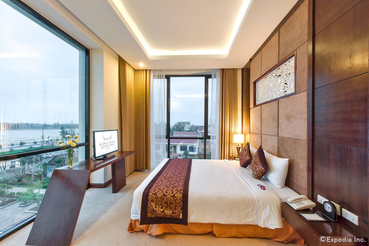 A hotel room with a large bed, a desk with a computer, a window with a view of the city, and a decorative wall panel.