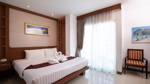 A spacious hotel room with a large bed, a wooden headboard, a chair, and a painting of a beach scene. - The Patong Center Hotel (Patong)