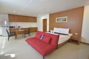 Quarto de hotel pequeno com cama, sofá vermelho, mesa e cozinha compacta.