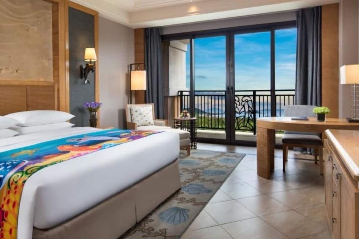 Une chambre d’hôtel avec un grand lit, un bureau, une chaise, un balcon avec vue et une grande fenêtre.