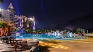 Une vue nocturne d’un hôtel avec une piscine, des palmiers et un grand bâtiment dont les fenêtres sont illuminées.