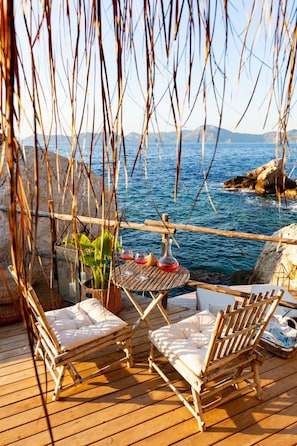 A wooden deck with two wicker chairs and a table, overlooking a body of water with rocks and hills in the background.