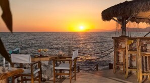 A sunset view from a wooden deck with a thatched roof, overlooking the sea.