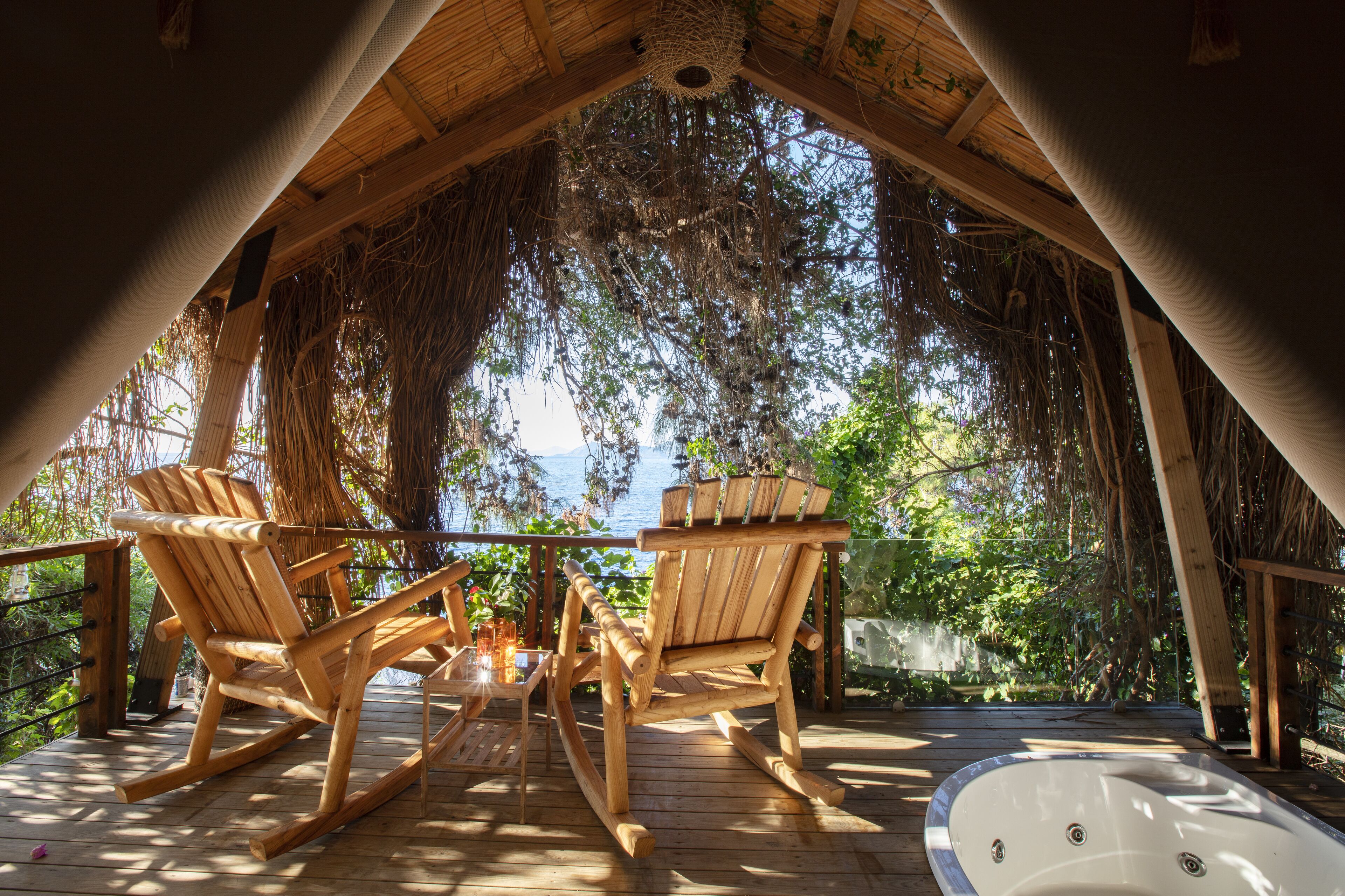 A wooden deck with rocking chairs and a hot tub, overlooking a lush garden and a body of water.