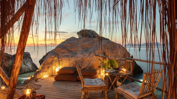 A wooden deck with a rustic seating area, including a cushioned chair and a wooden table with a candle, overlooking a rocky coastline at sunset. - Perdue Hotel (Fethiye)