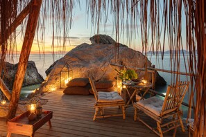 A wooden deck with a rustic seating area, including a cushioned chair and a wooden table with a candle, overlooking a rocky coastline at sunset. - Perdue Hotel (Fethiye)