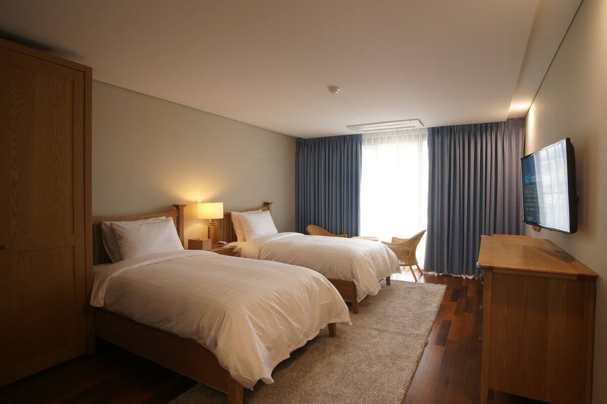 A hotel room with two beds, a wooden wardrobe, a desk, a television, and a large window with curtains.
