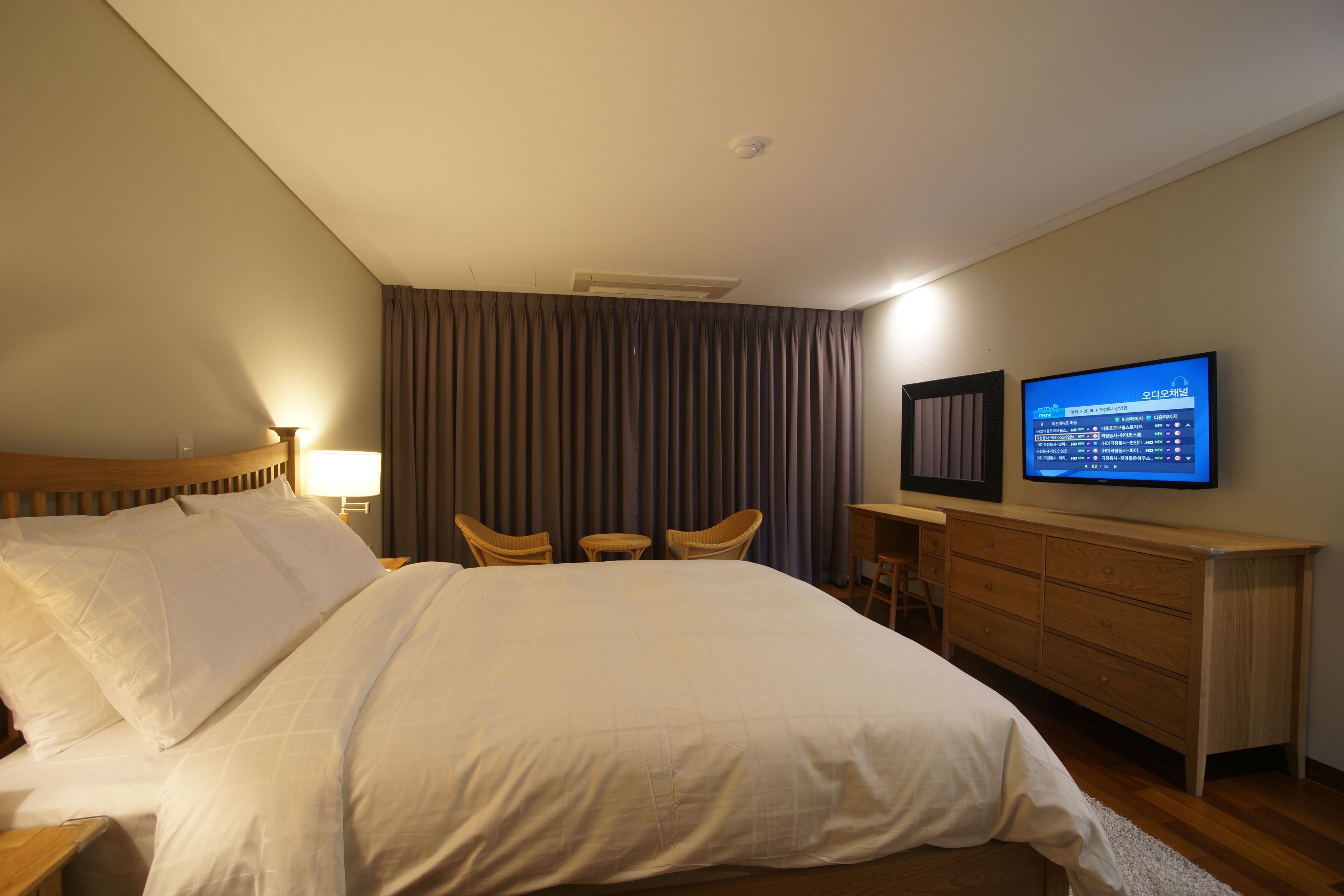 Quarto de hotel com uma cama grande, televisão de tela plana na parede, cômoda de madeira e uma escrivaninha com duas cadeiras.