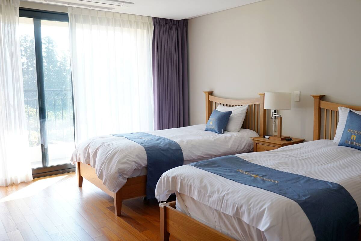 A hotel room with two beds, a large window with curtains, a wooden floor, and a bedside table with a lamp.