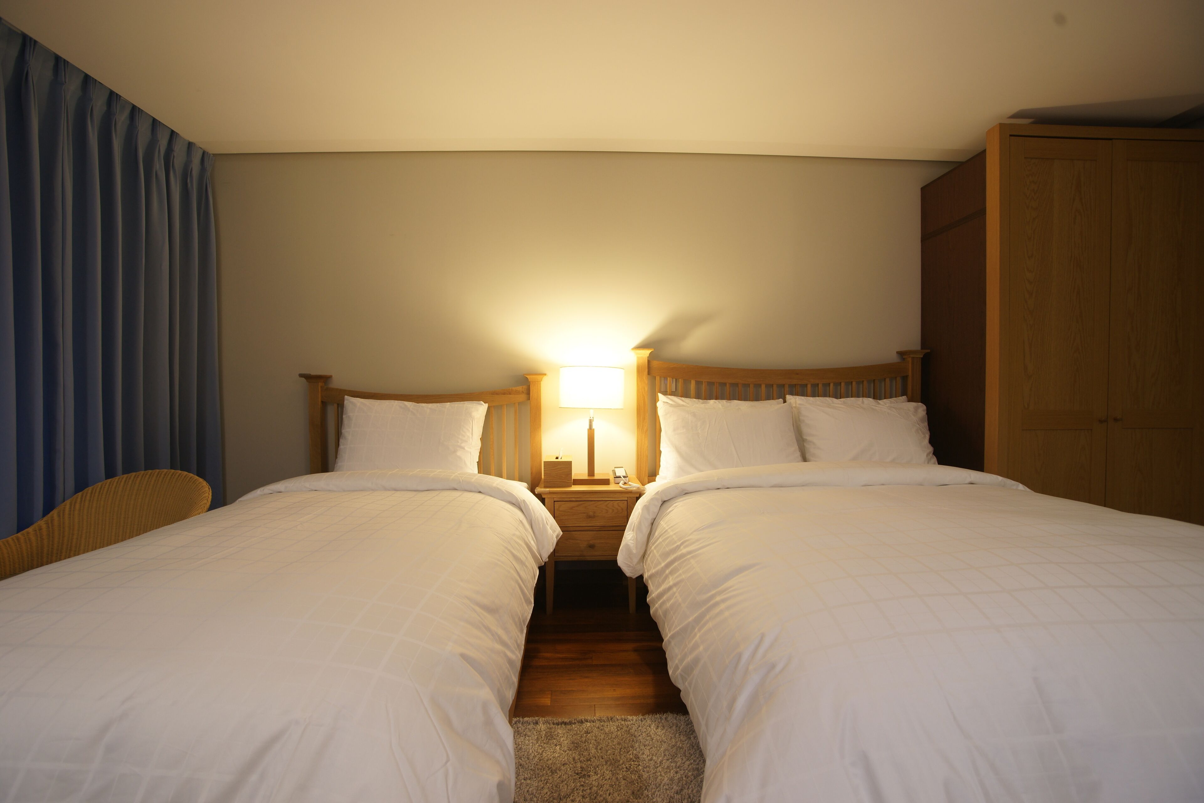 Une chambre d’hôtel avec deux lits simples, une armoire en bois, une table de chevet avec une lampe et un rideau bleu.