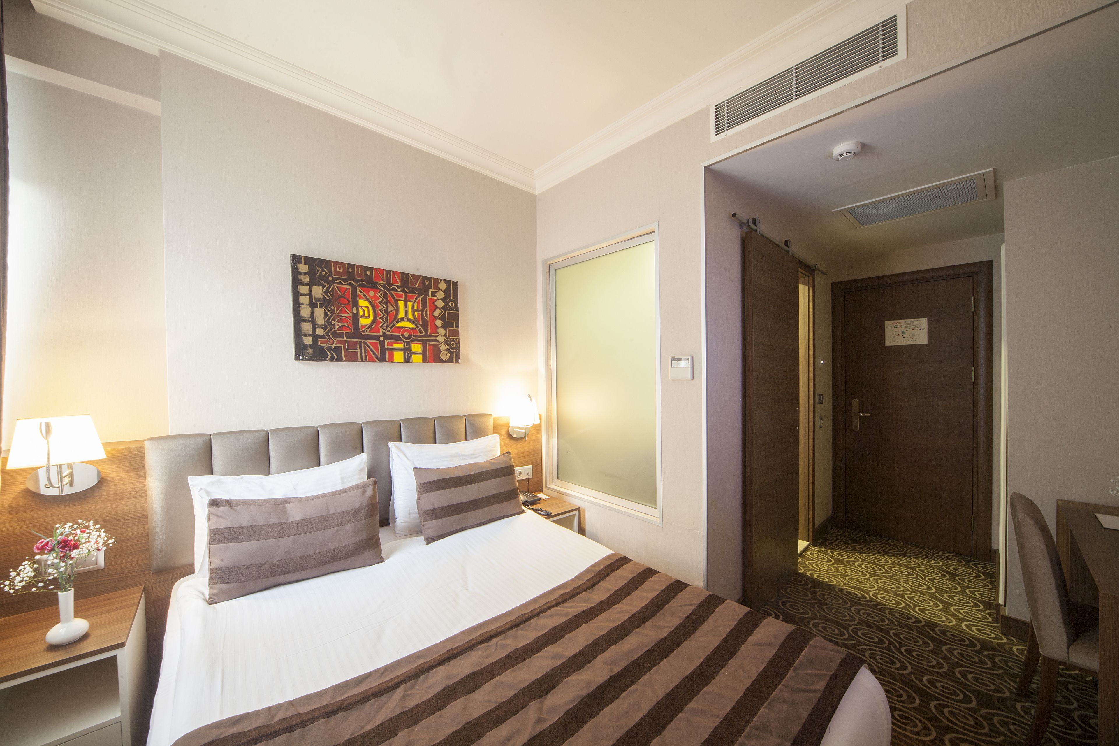 Une chambre d’hôtel avec un lit, une table de chevet, une lampe, un tableau au mur et une porte donnant sur une autre pièce.