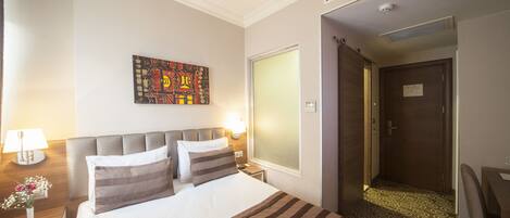 Habitación de hotel con cama, mesita de noche, lámpara, un cuadro en la pared y una puerta que da a otra habitación.