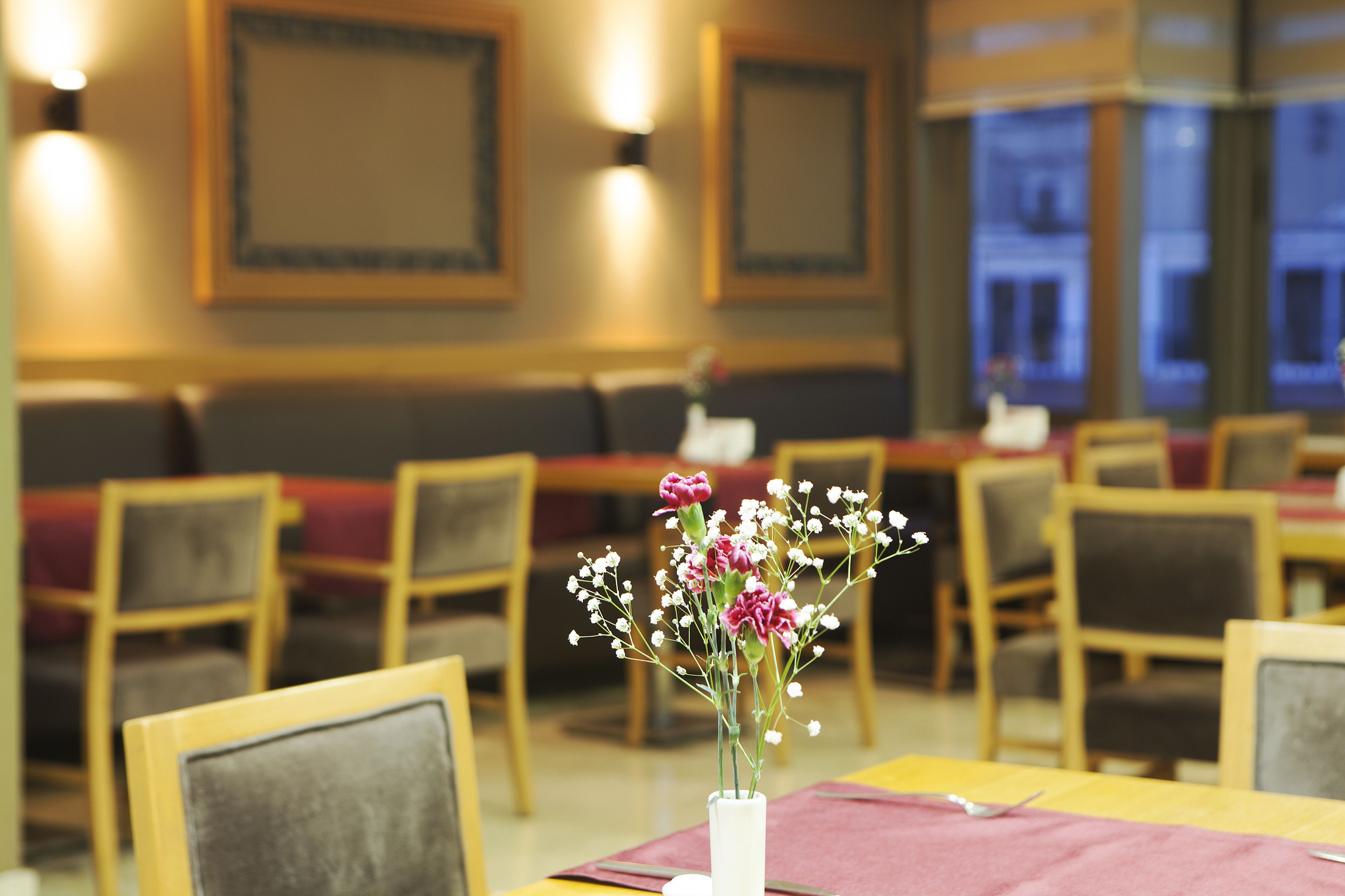 A restaurant interior with wooden chairs, a table with a vase of flowers, and framed artwork on the walls.