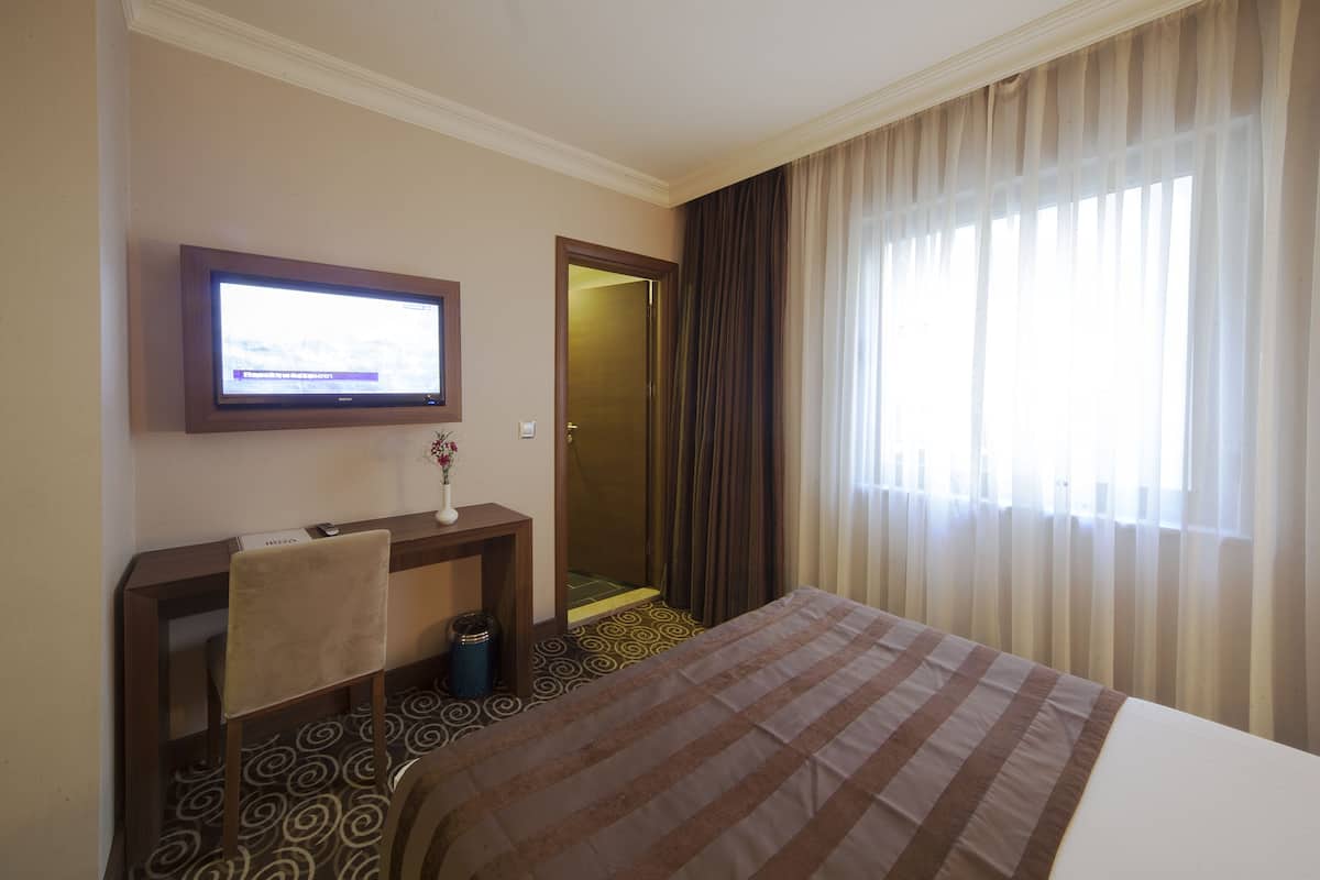 Habitación de hotel con cama, escritorio con silla, televisor en la pared y ventana con cortinas.