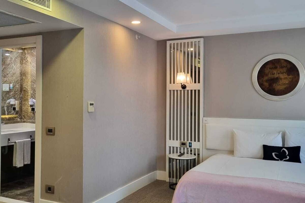 A hotel room with a bed, a bedside table, a wall-mounted lamp, a mirror, and a bathroom visible through an open door.