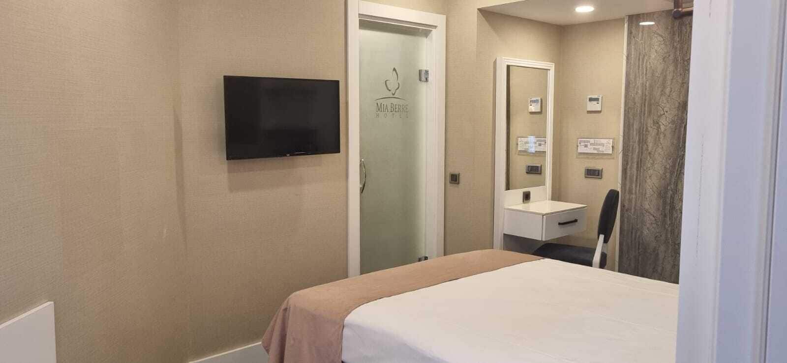 A hotel room with a bed, a television mounted on the wall, a bathroom with a mirror, and a door with a logo.