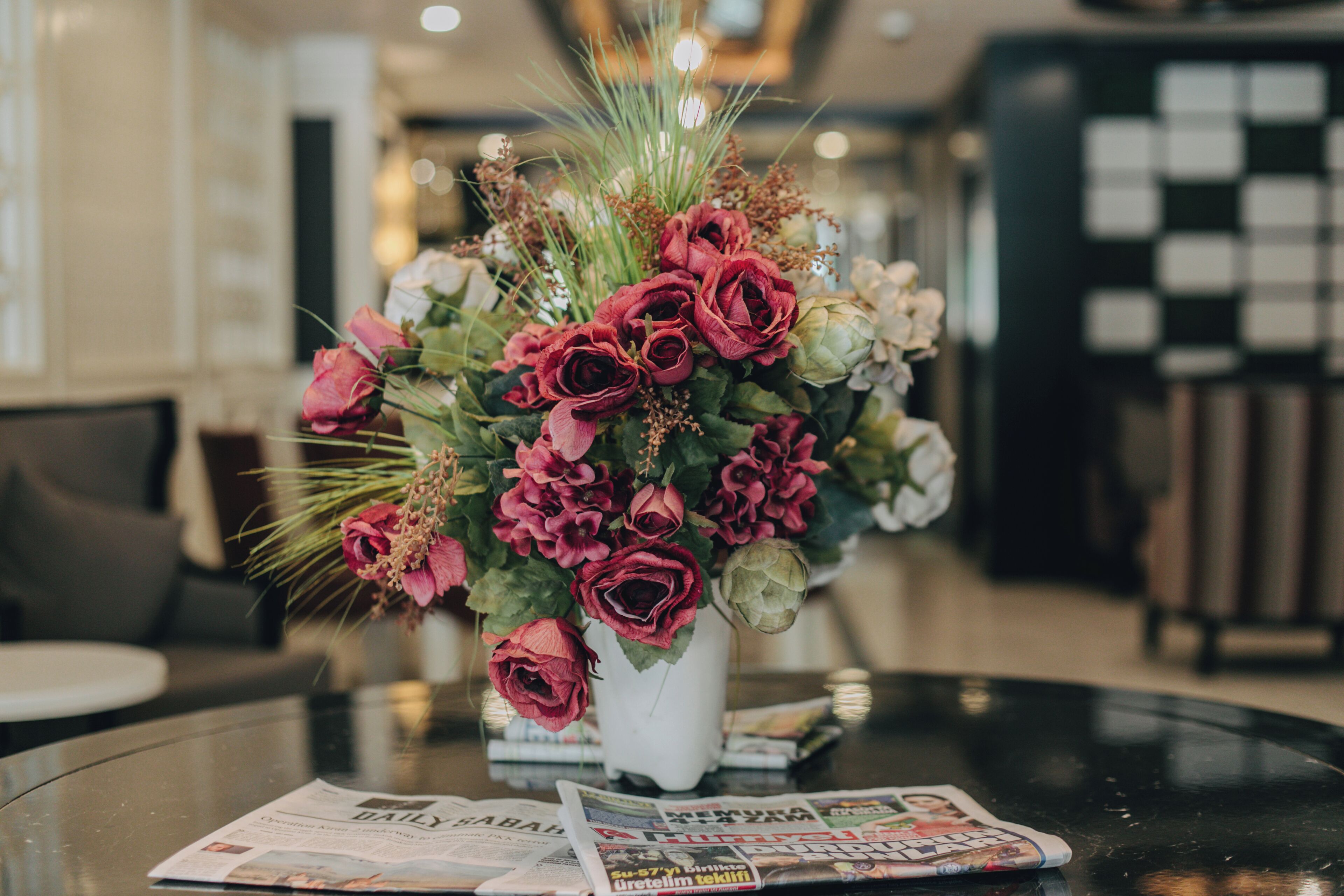 A table with a bouquet of flowers in a white vase and some newspapers.