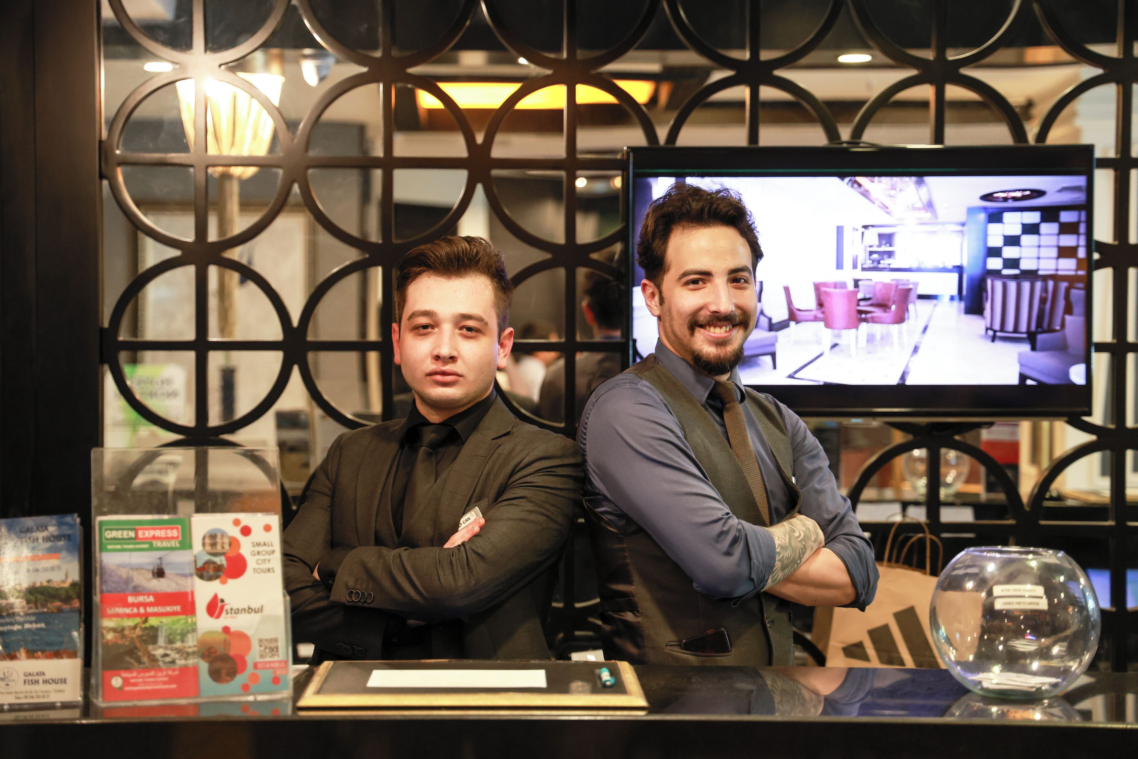 Two men seated at a reception desk with a television screen displaying a room interior.
