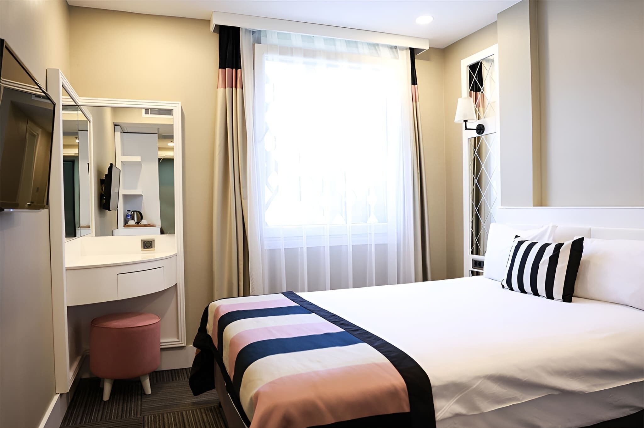 Una habitación de hotel con cama, tocador con espejo, un pequeño taburete, ventana con cortinas y una lámpara de pared.