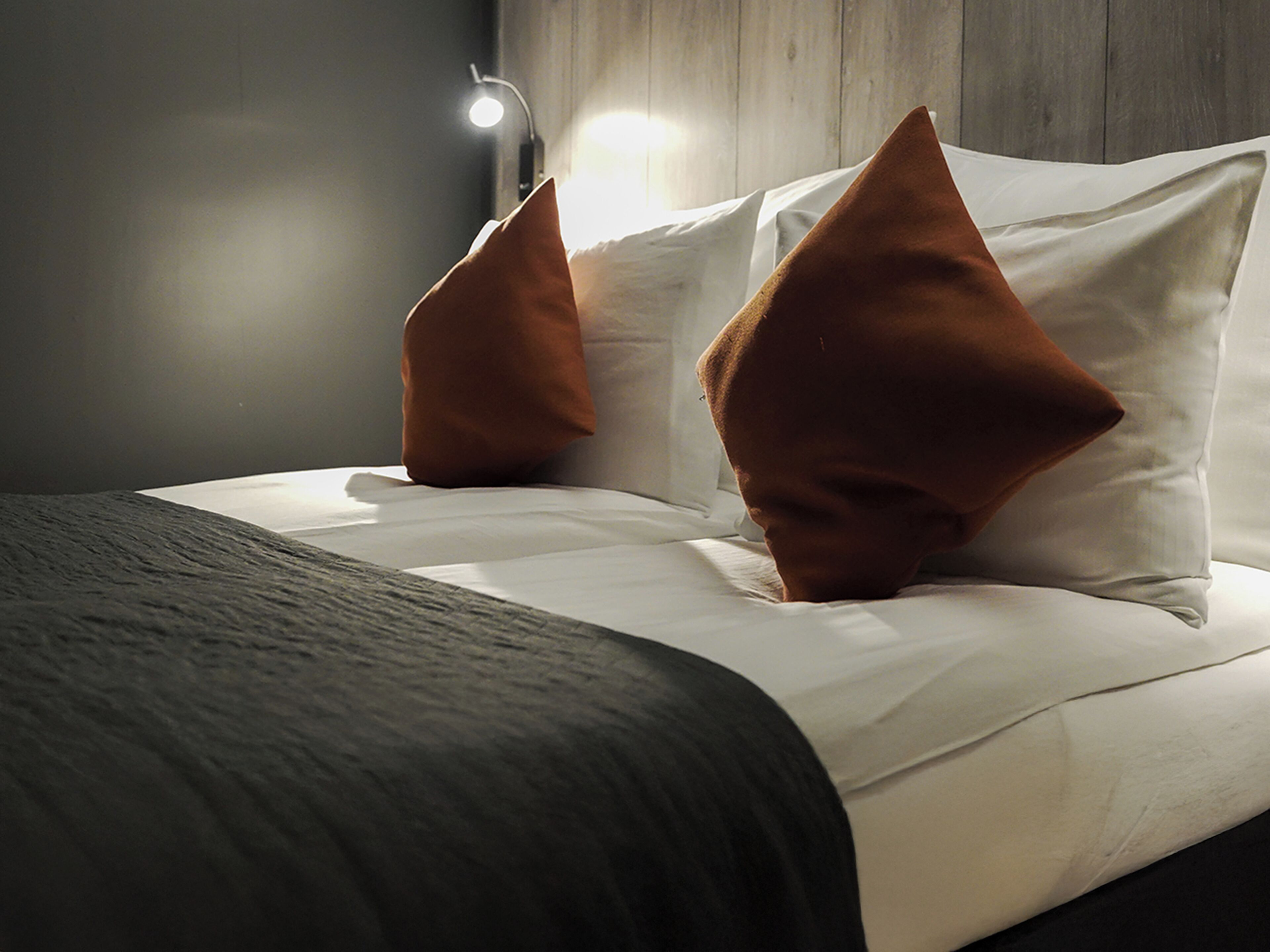 Una cama bien hecha con sábanas blancas y dos almohadas marrones, una lámpara de noche y un cabecero de madera.