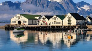 A waterfront building with a green roof, a dock with boats, and a mountain range in the background. - Arctic Panorama Lodge (Skjervoy)