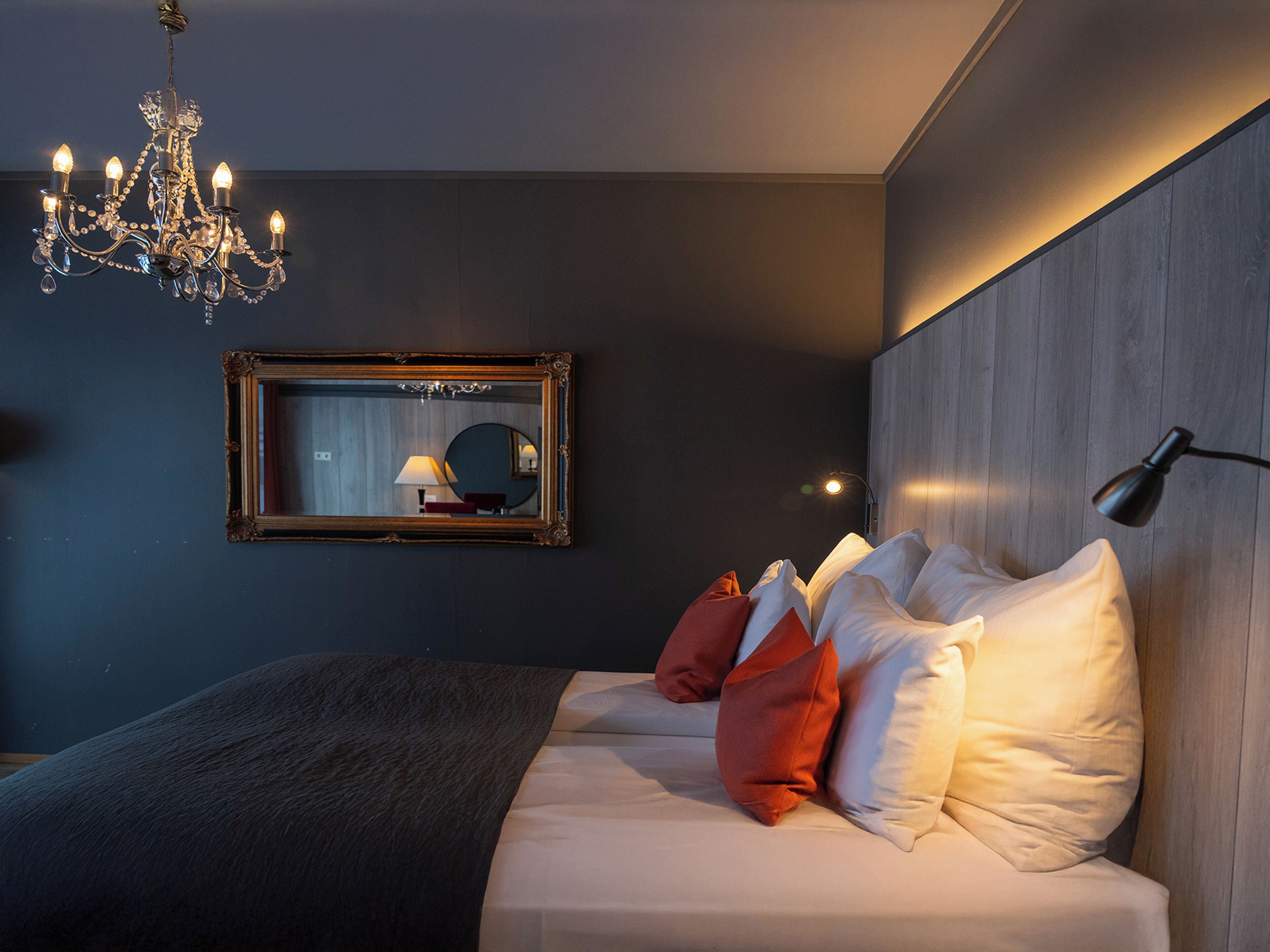 A modern bedroom with a large bed, a chandelier, a mirror, and a bedside lamp.